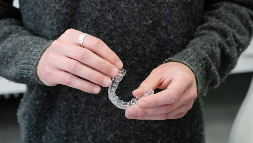 Harmaaseen neuleeseen pukeutunut henkilö pitää käsissään läpinäkyviä Invisalign-oikomiskalvoja.